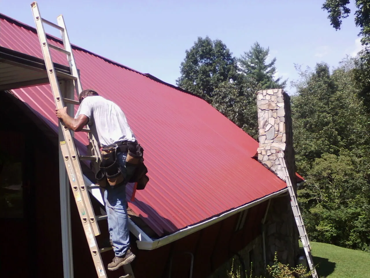 Roofer on a ladder beside a red metal roof and stone chimney — Soffit & Fascia Repair by Roofing Brothers in Swisher